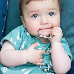 A baby with blue eyes and light brown hair is wearing a green onesie with bird prints. They are holding a YUMMIKEYS Yummirings - Stainless Steel Teething Ring and biting on it to soothe their teething gums. The background includes blue and teal cushions.