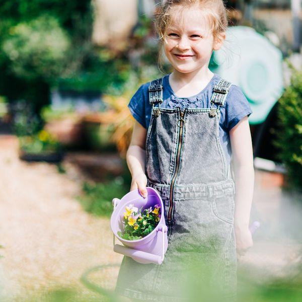 Child in dungarees holding a purple bucket with plants in it.