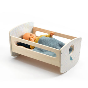 A baby doll, snugly wrapped in a blue and green blanket, rests peacefully in the Djeco Rocking Bed Cradle, featuring a natural wood finish and a charming heart-shaped cutout on the headboard. The doll is dressed in a yellow top.