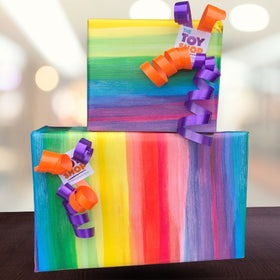 Two gift boxes wrapped in the colorful designs from Wrapped's Wrapping Service are placed on a dark wooden surface. Each package is adorned with orange and purple curly ribbons, giving it an extra-special touch. A small tag that reads "The Toy Shop" is attached to both ribbons, ideal for adding a personal message. The background is softly blurred.