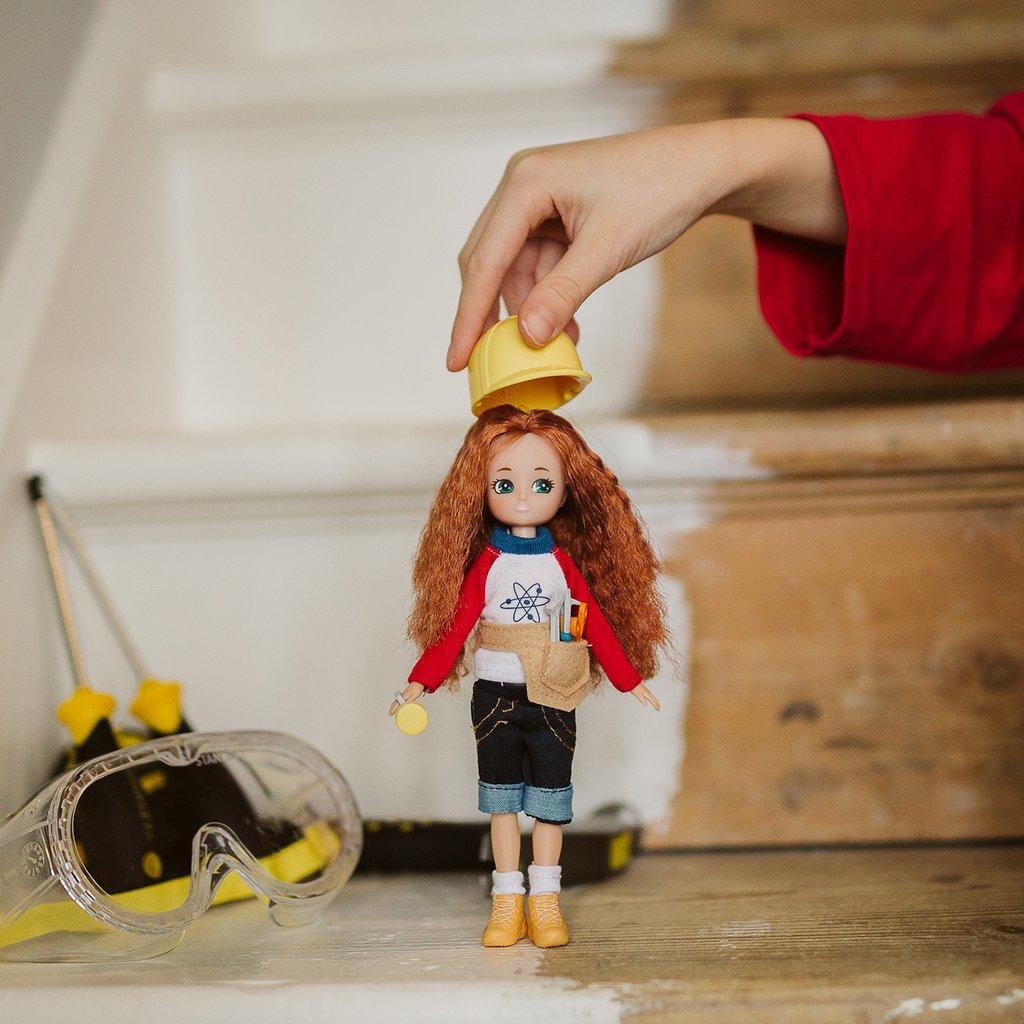 Lottie Inventor Doll with long red curly hair stands at the foot of wooden and partially white painted stairs as a child's hand places a doll's yellow hard hat on her head.