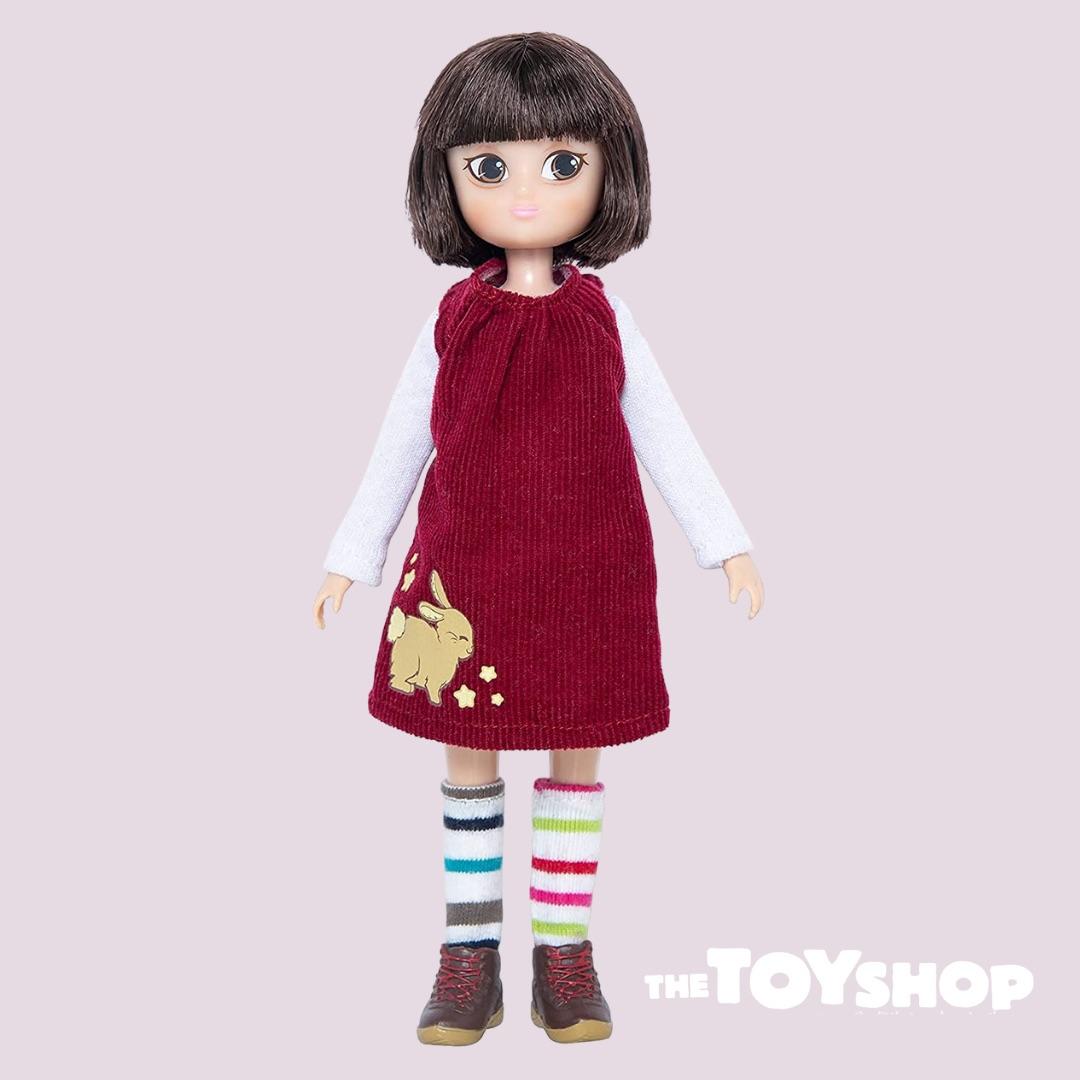 Child's doll with dark fringed hair and a deep red dress over a long-sleeve white t-shirt and wearing odd stripy socks and ankle boots.