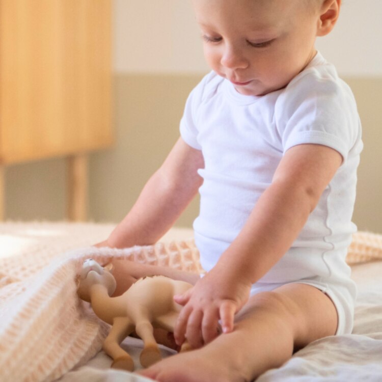 A baby in a white onesie sits on a bed, playing with Al' Thir the Companion toy by Sophie La Giraffe, made from natural rubber. The softly lit room has a wooden nightstand partially visible in the background.