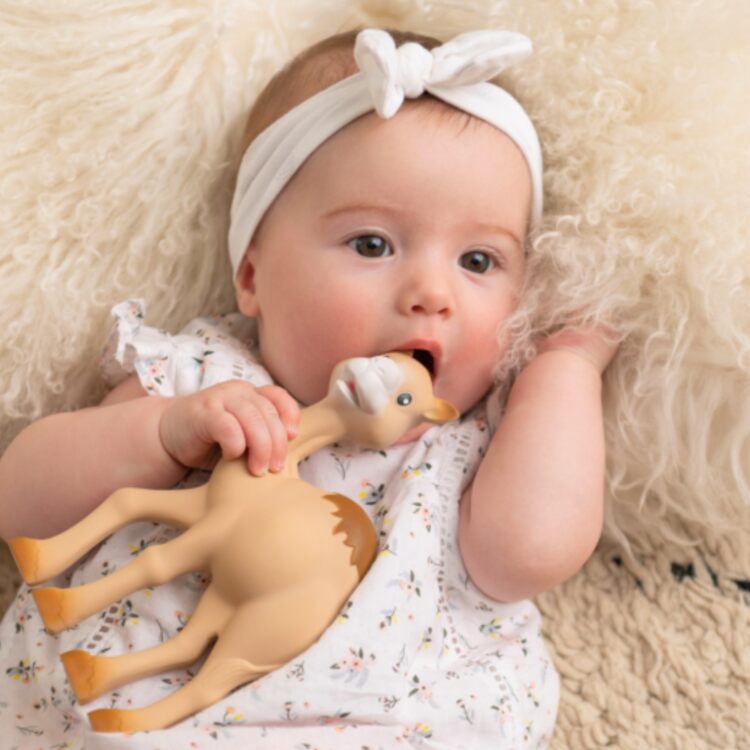 A baby wearing a patterned outfit and white headband lies on a textured blanket, holding Al' Thir the Companion by Sophie La Giraffe, a brown natural rubber toy resembling a giraffe. Gazing curiously at the camera, the toy provides teething relief and comfort.