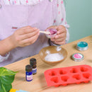 A person wearing a white shirt and purple overalls uses the KISS NATURALS DIY Natural Bath Bomb Kit, mixing a pink substance in a jar. The table features all-natural ingredients, including white powder, essential oils, and a red heart mold. A green plant leaf adds nature on the left.