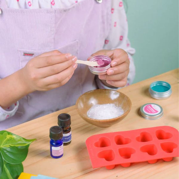A person wearing a white shirt and purple overalls uses the KISS NATURALS DIY Natural Bath Bomb Kit, mixing a pink substance in a jar. The table features all-natural ingredients, including white powder, essential oils, and a red heart mold. A green plant leaf adds nature on the left.