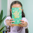 A child happily holds a green "DIY Natural Bath Bomb Kit" from KISS NATURALS, showcasing colorful designs and bilingual text. Ideal for tweens and teens, it uses all-natural ingredients. A softly blurred plant is seen against a light wall in the background.