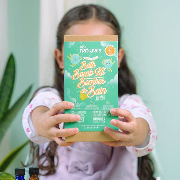 A child happily holds a green "DIY Natural Bath Bomb Kit" from KISS NATURALS, showcasing colorful designs and bilingual text. Ideal for tweens and teens, it uses all-natural ingredients. A softly blurred plant is seen against a light wall in the background.