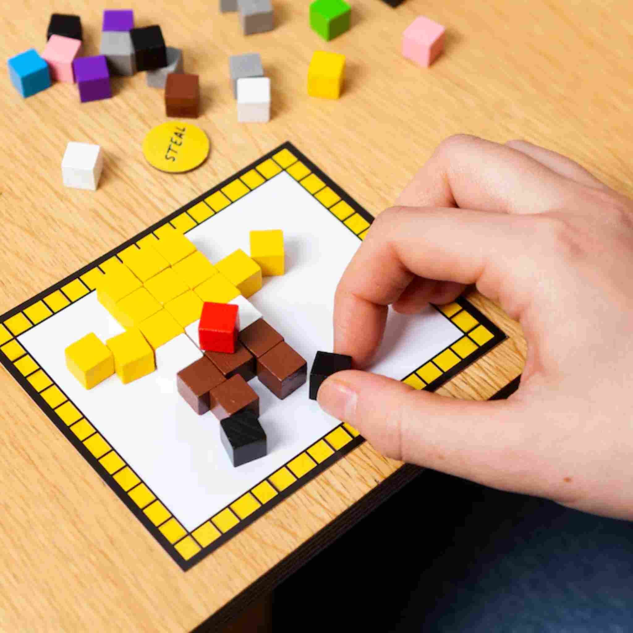 A hand places a black cube on a card with a yellow border, creating a pixelated figure for Big Potato's Block Party block-building game. The cubes, available in yellow, brown, and black, are scattered across the wooden surface like pieces from Block Party challenges.