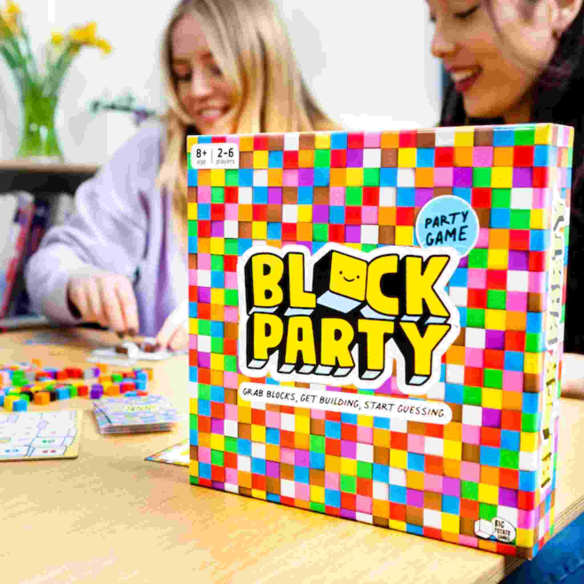 Two individuals are seated at a table, deeply engaged in an imaginative block-themed board game named "Block Party" by Big Potato. Featuring the tagline, "Grab blocks, get building," this thrilling game invites players to dive into the world of guessing and construction. Surrounded by a vibrant room adorned with flowers, it creates the ideal setting for their entertaining experience.