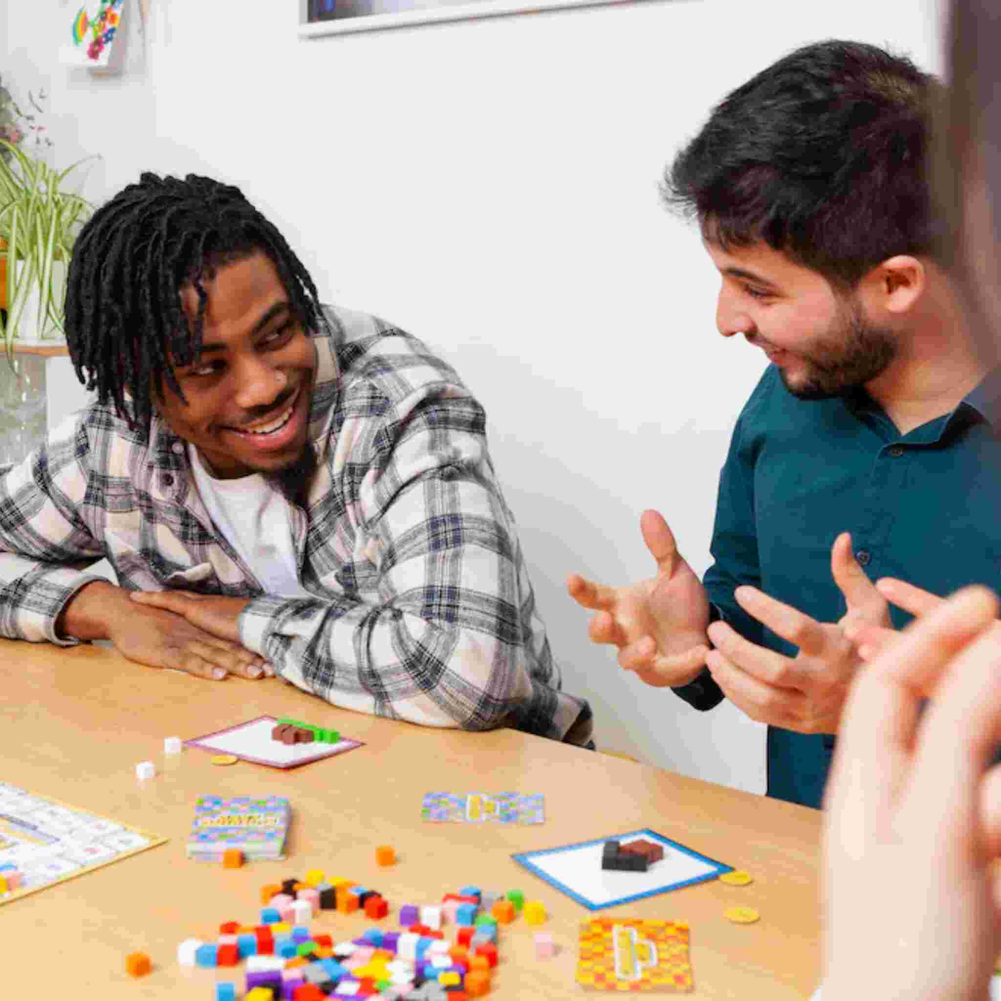 Two people sit at a table, deeply engaged in the vibrant Block Party board game by Big Potato, smiling and chatting. The colorful pieces are spread across the table, enhancing the excitement. A small plant is visible in the background, adding a touch of nature to their spirited Block Party challenges.