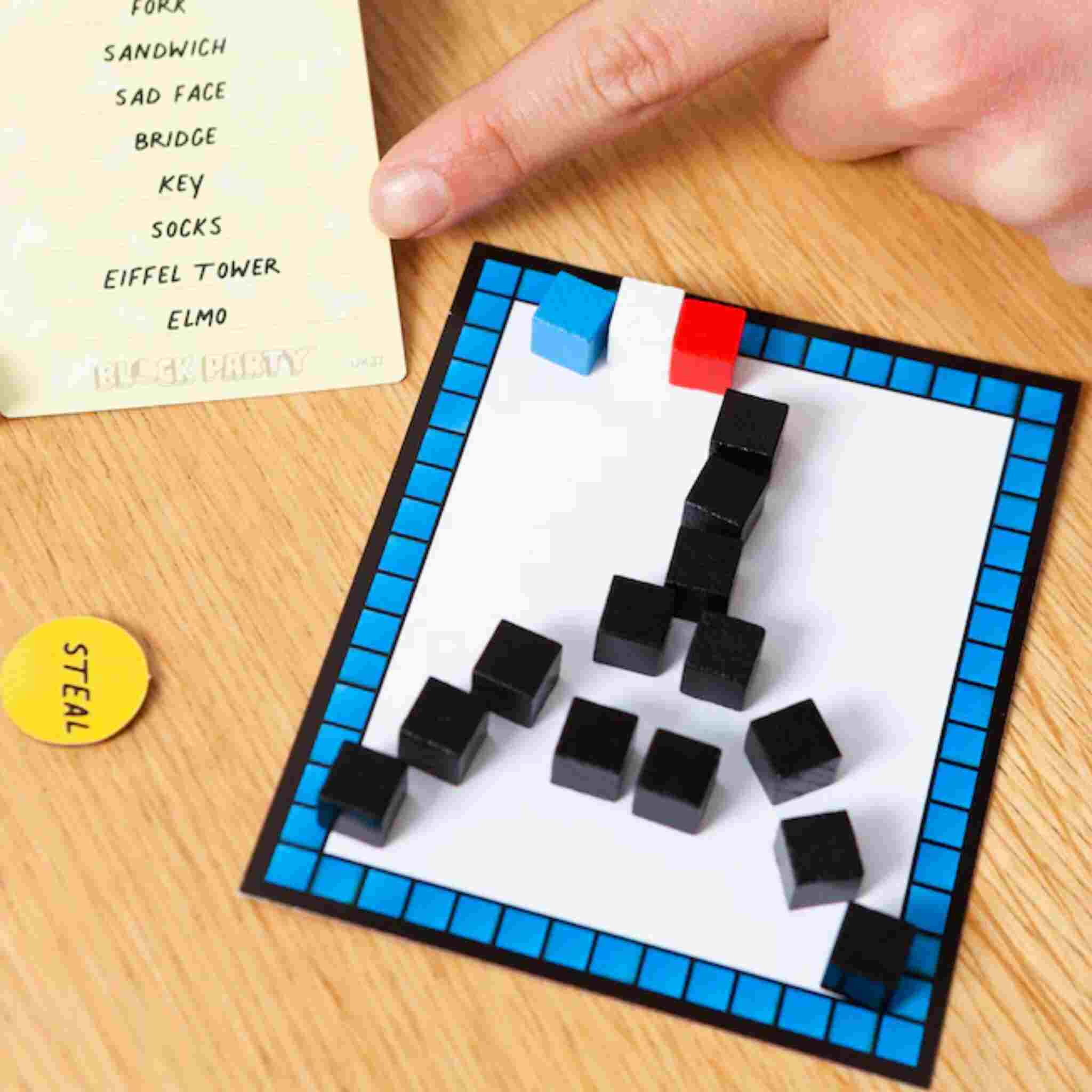 A hand points at a drawing made of black blocks resembling the Eiffel Tower on a small board with a blue border, similar to the challenging Block Party game by Big Potato. On the wooden table nearby, there's a list of words and a yellow "steal" token, ready to add excitement to your Block Party challenges.