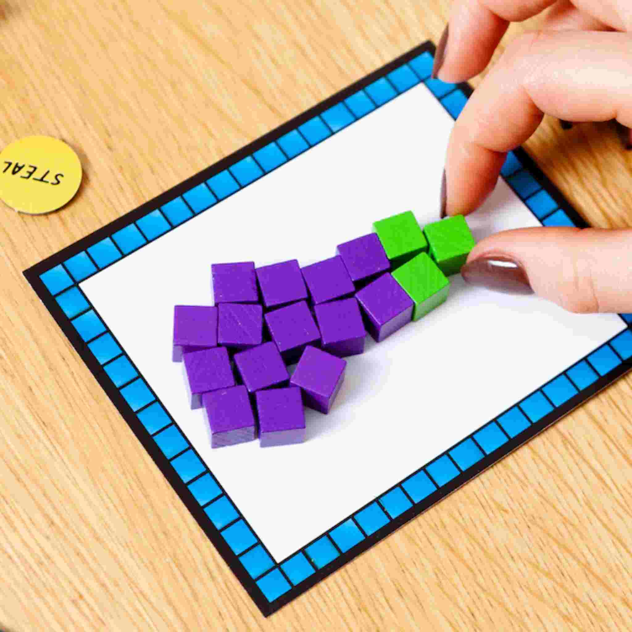 A hand arranges colorful wooden cubes on a framed white card, evoking memories of a block-building game. Purple cubes are crafted into the shape of a bunch of grapes, with three green cubes as the stem. A yellow circular token featuring "STEAL" suggests challenges from the Block Party game by Big Potato on the wooden surface.