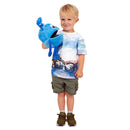 A young child with blonde hair holds a large Blue Baby Monster Puppet by THE PUPPET COMPANY, resembling a fish. The child wears a bicycle-printed T-shirt, olive green shorts, and brown sneakers, smiling with sparkling eyes and looking slightly to the side with a squeaky voice.