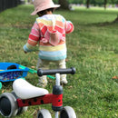 In a vibrant, summery park, a toddler in a colorful striped hoodie and pink hat stands on the grass beside the Boldcube Baby Balance Bike - Betty Ladybird and a red tricycle, making it an idyllic setting for selecting a jungle-themed first birthday gift.