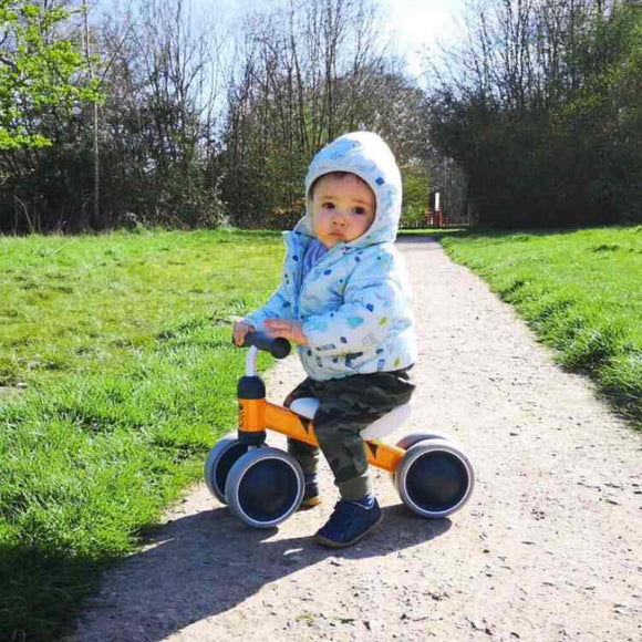 A toddler in a blue jacket and hood rides the orange Boldcube Baby Balance Bike - Benny Tiger down a dirt path, surrounded by lush green grass and trees, reminiscent of an adventurous jungle-themed first birthday gift trail. The sun shines brightly, making it a perfect day.