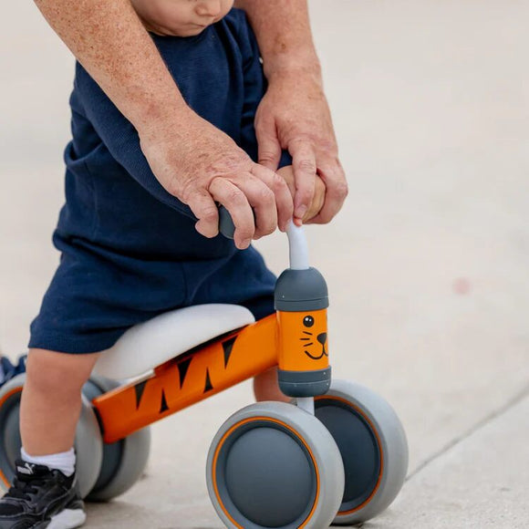A toddler in navy blue shorts holds the handlebars of a Boldcube Baby Balance Bike - Benny Tiger, featuring playful animal designs, perfect as a jungle-themed first birthday gift. An adult gently guides them on the concrete, introducing them to baby bikes.