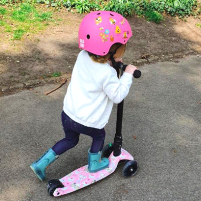 A child wearing a vibrant pink BOLDCUBE Kids Helmet, designed from impact-resistant materials, sports a white jacket and blue boots while riding a matching pink scooter along a park's paved path, surrounded by lush grass and dirt patches.