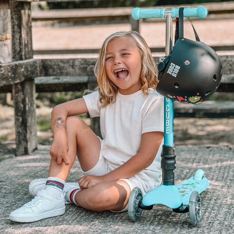 Blonde haired child wearing white sitting next to turquoise scooter with black helmet hanging from handle.
