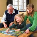 A young girl, an older man, and a woman sit on a sofa, playing a board game in a bright room with large windows. The smiling girl is as excited as if she were piecing together the Cobble Hill "Call of the Wild" jigsaw puzzle as she moves her game piece on the colorful board.