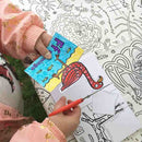 A child, immersed in a children's craft activity with the eggnogg Card Book - Pop-up Animals, skillfully maneuvers scissors to cut out a colorful flamingo drawing amidst clouds and fish, set against a detailed black-and-white illustrated tablecloth that inspires imaginative pop-up cards.