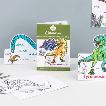 A table showcases eggnogg's "Card Book – Pop-up Dinosaurs," featuring vibrant pop-up cards with a Tyrannosaurus and outlined dinosaurs, inviting creativity with partially colored bright hues.