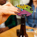 A hand holds an eye-catching, colorful circular disk with a wavy pattern from Big Potato's "Cards vs Gravity" set and places it on top of a brown bottle. In the background, people sit at a table, slightly out of focus.