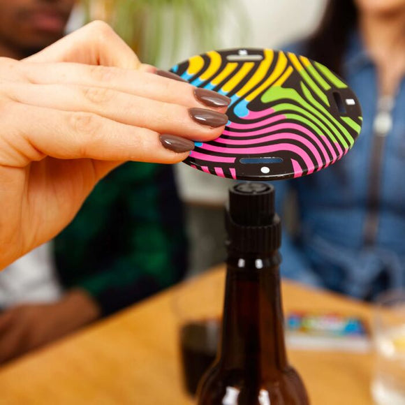 A hand holds an eye-catching, colorful circular disk with a wavy pattern from Big Potato's "Cards vs Gravity" set and places it on top of a brown bottle. In the background, people sit at a table, slightly out of focus.