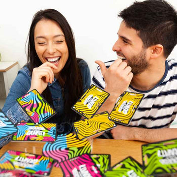 A smiling man and woman sit at a table, laughter echoing as they enjoy a game of Cards vs Gravity by Big Potato, with colorful, waterproof cards scattered about—like "Jenga in your pocket," these lively cards promise endless fun without the worry of spills.