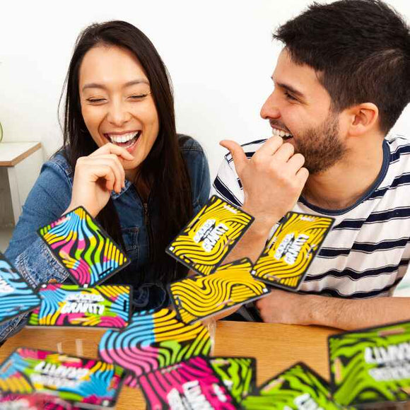 A smiling man and woman sit at a table, laughter echoing as they enjoy a game of Cards vs Gravity by Big Potato, with colorful, waterproof cards scattered about—like "Jenga in your pocket," these lively cards promise endless fun without the worry of spills.