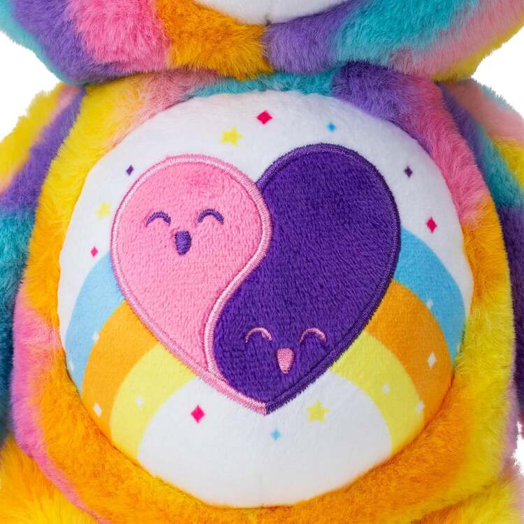 Close-up of the Care Bears Eco-friendly Friends Forever (35cm) by Basic Fun, a vibrant plush toy with a rainbow pattern and an embroidered belly design featuring overlapping pink and purple hearts surrounded by small stars and a rainbow.