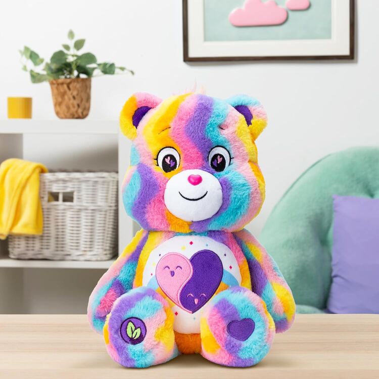 A Care Bears Eco-friendly Friends Forever plush by Basic Fun, featuring a rainbow pattern and large embroidered eyes, sits on a wooden surface. The bear has a heart motif with two smaller hearts on its belly, while the background includes a green chair, shelf with basket, and wall art.