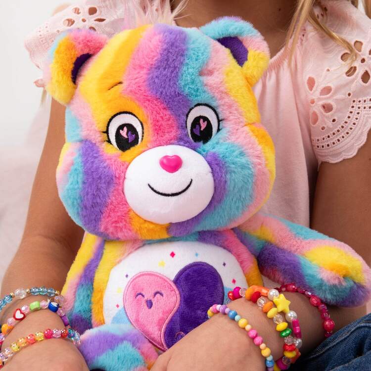 A child is holding the Care Bears Eco-friendly Friends Forever bear (35cm) by Basic Fun, adorned with rainbow fur and heart designs. They wear a pink lace-detailed top and beaded bracelets, capturing the vibrant essence of Care Bears.