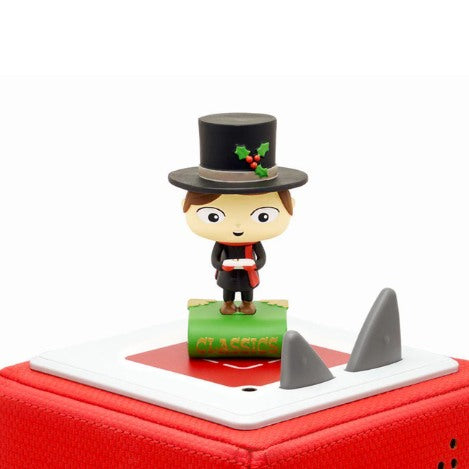 A small figurine from Tonies' "Tonie - A Christmas Carol and other Classic Stories" stands on a red square base, capturing the essence of timeless literature. The figure, wearing a top hat adorned with holly and holding a scroll, is set on a base labeled "CLASSICS" and has two triangular protrusions on the side.
