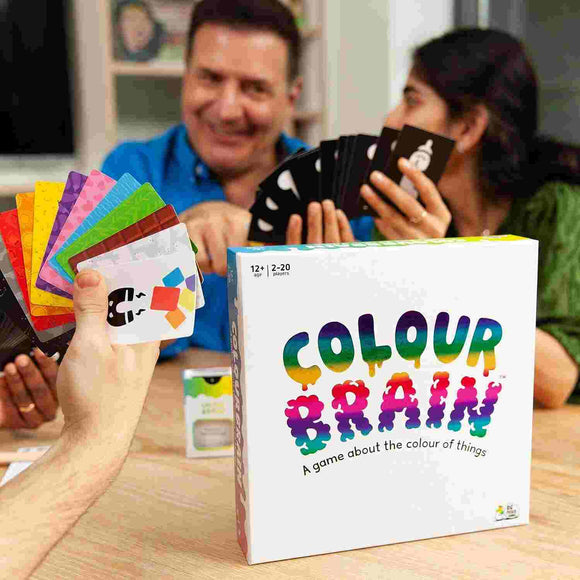 A group of people engaged in fun plays Big Potato's family game, "Colour Brain," with one holding colorful cards to answer color questions. The game's box is prominently displayed in the foreground as everyone enjoys themselves.