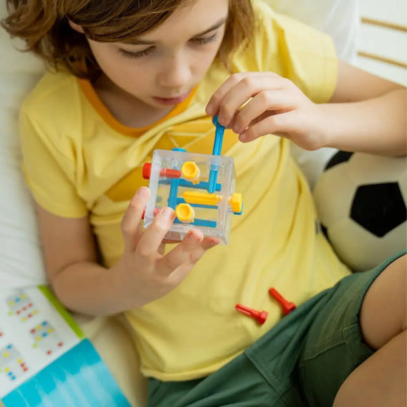 A child in a yellow shirt and green shorts focuses on a Criss Cross Cube by Smart Games, accompanied by a black-and-white soccer ball and guided by an instruction booklet partially in view. The transparent 3D puzzle features blue and yellow elements for engaging problem-solving fun.