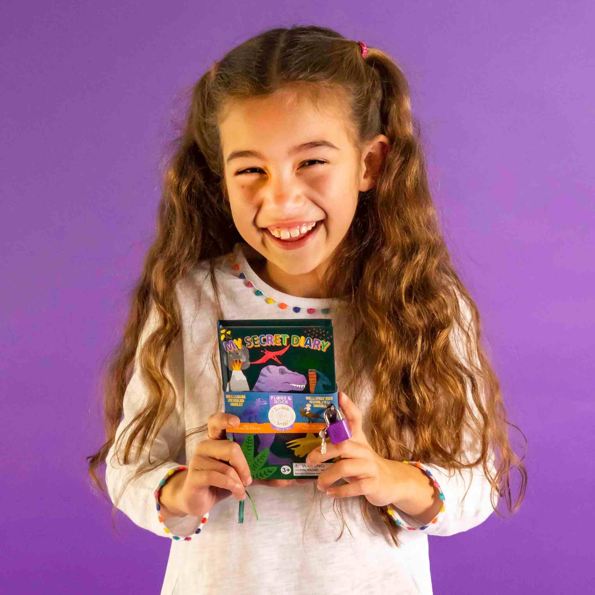 A smiling girl with long hair in pigtails holds the Dino My Scented Secret Diary by Floss & Rock against a purple background. She's wearing a white shirt with colorful accents on the cuffs, perfectly complementing the vibrant scene.