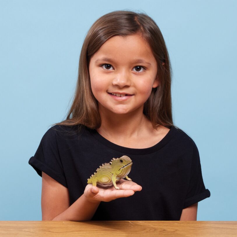 A girl with long brown hair, wearing a black t-shirt, smiles while holding a small EUGY Tuatara in her hand. She is seated at a wooden table against an eco-friendly, light blue background that enhances the charm of their collectable moment.