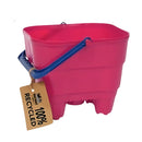 The Crazy Coast Eco 100% Recycled Castle Bucket is a bright pink, rectangular container with a plant symbol, blue handle, brown "100% Recycled" tag, small top hook, and raised base for easy handling—ideal as an eco-friendly children's product.