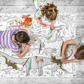 Three children are engrossed in a craft activity, coloring an eggnogg Giant Poster/Tablecloth – Dinosaurs on the wooden floor. With an assortment of markers and toy dinosaurs, they animate the detailed patterns within the poster's many dinosaur designs.