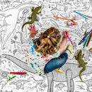 A blonde-haired child is coloring an eggnogg Giant Poster / Tablecloth – Dinosaurs on the floor, surrounded by vibrant pencils and toy dinosaurs, while wearing a colorful splatter-patterned shirt and jeans, bringing prehistoric wonders to life.