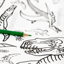 A green colored pencil sits on the Giant Poster / Tablecloth – Dinosaurs by eggnogg, a white coloring-in poster featuring outlined dinosaur drawings such as a large head and other prehistoric creatures—ideal for an enjoyable craft activity.