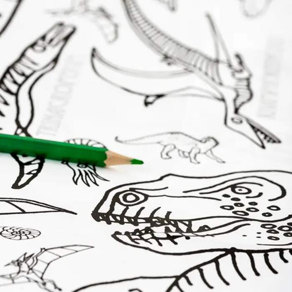 A green colored pencil sits on the Giant Poster / Tablecloth – Dinosaurs by eggnogg, a white coloring-in poster featuring outlined dinosaur drawings such as a large head and other prehistoric creatures—ideal for an enjoyable craft activity.