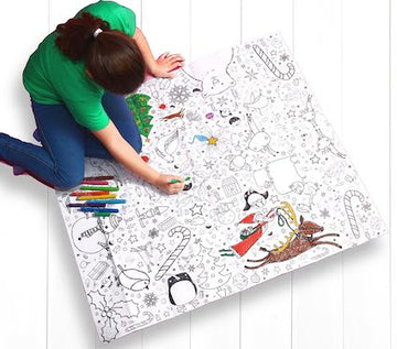 A child in a green shirt is deeply focused on coloring an Eggnog Christmas Colour-In Giant Poster / Tablecloth, which features holiday-themed designs filled with candy canes, animals, and stars. Colorful markers are scattered around on the white floor as the Christmas coloring unfolds.