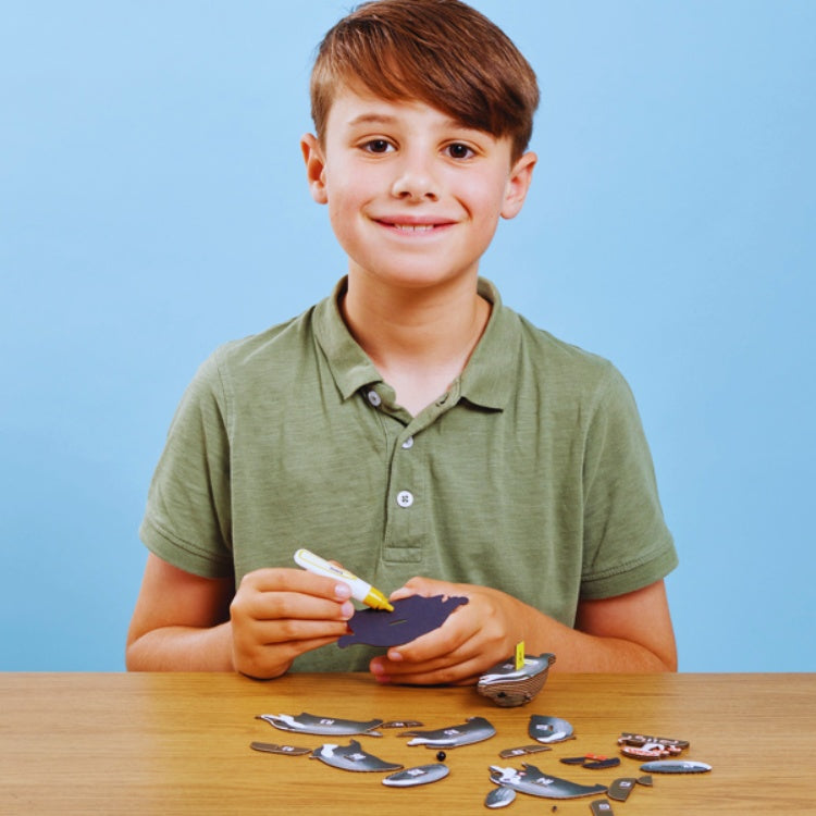 A young boy with brown hair in a green shirt smiles as he assembles an EUGY whale-shaped puzzle, crafted from sustainable materials, at a wooden table. A blue background provides the perfect backdrop for his new collection of EUGY toy figures.