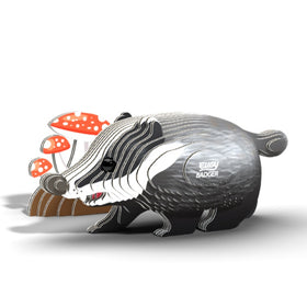 The EUGY Badger, a stylized, cartoonish figurine crafted from sustainable materials, stands beside red and white mushrooms on a plain white background. As part of the EUGY toy figures collection, it features "Busy Badger" on its side with distinct black, white, and gray stripes.