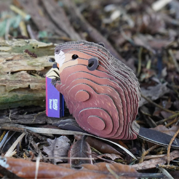 The EUGY Eugy Beaver is a collectible, eco-friendly paper-craft puzzle model featuring a beaver holding a purple notebook and sitting among twigs, leaves, and tree bark.