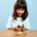 A young girl with long dark hair wearing a light blue shirt sits at a table against a blue backdrop, assembling an EUGY Bat with eco-friendly cardboard cutouts. A glue stick is visible on the table as she focuses intently and smiles, enjoying her craft with sustainable materials from EUGY.