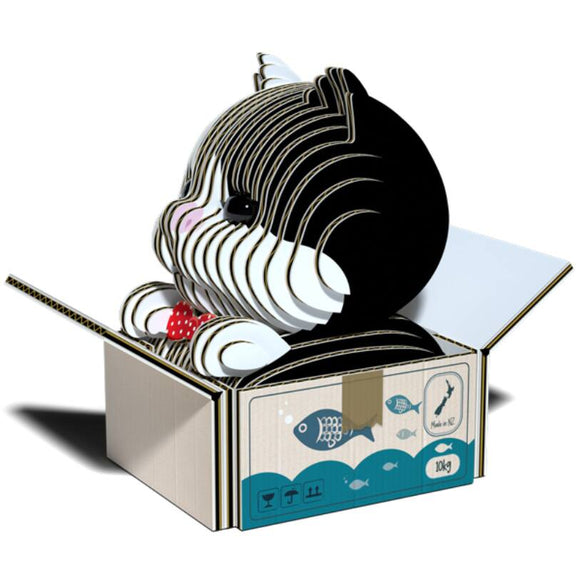 The Eugy Cat - Tuxedo by EUGY is an eco-friendly 3D cardboard sculpture crafted with horizontal layers, elegantly displayed inside an open box. It showcases a charming black and white pattern with a red bow tie, making it a delightful addition to any collection. The box is embellished with fish illustrations and other whimsical symbols.