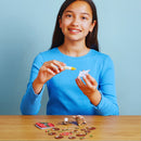 A girl in a blue sweater is crafting at a table. She holds a glue pen and a small paper cutout, resembling an EUGY Christmas Elf. Various decorative pieces, including parts of EUGY toy figures, are spread out in front of her against a light blue background.